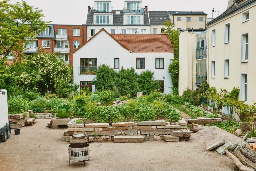 Der Marmorgarten 2023: zu sehen ist ein voll bewachsenes Hochbeet und ein Pyrolyseofen von Kon-Tiki. Im Hintergrund das 2 stöckige Nachbarhaus. Fotograf: Robert Schlossnickel.